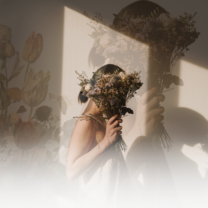 A person holds dried flowers in front of their face, casting shadows on a floral wallpaper backdrop.