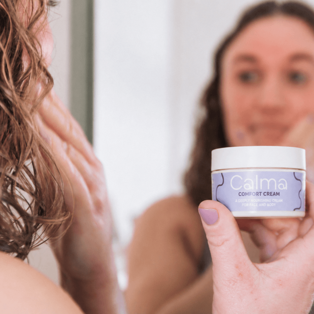 A person holding a jar of Calma Comfort Cream in front of a mirror, with their hand and hair visible and their reflection blurred in the background.