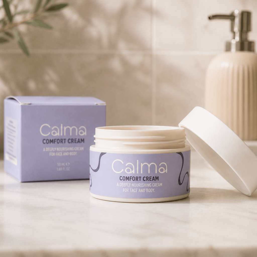 An open jar of Calma Comfort Cream sits on a marble countertop with its matching lavender box behind it and a cream-colored pump bottle blurred in the background.