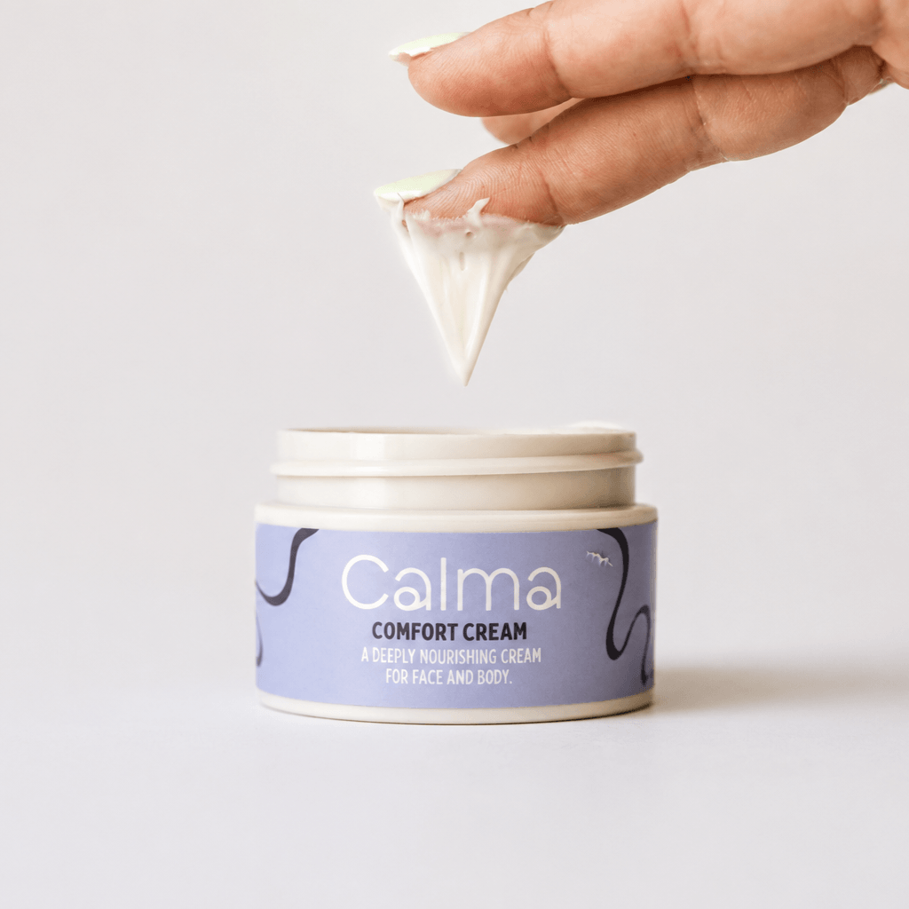 A hand holding a small pointed piece of white cream above an open jar of Calma Comfort Cream on a clean white background.