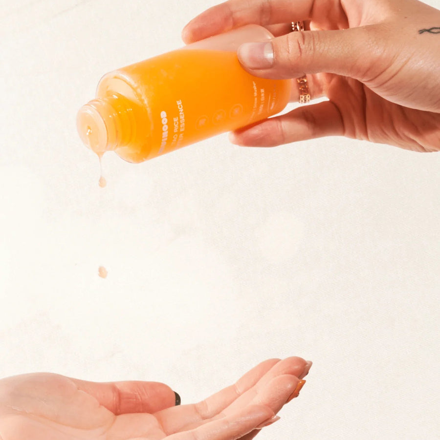 Hand holding an orange bottle with a white label, squeezing liquid onto another hand against a light background