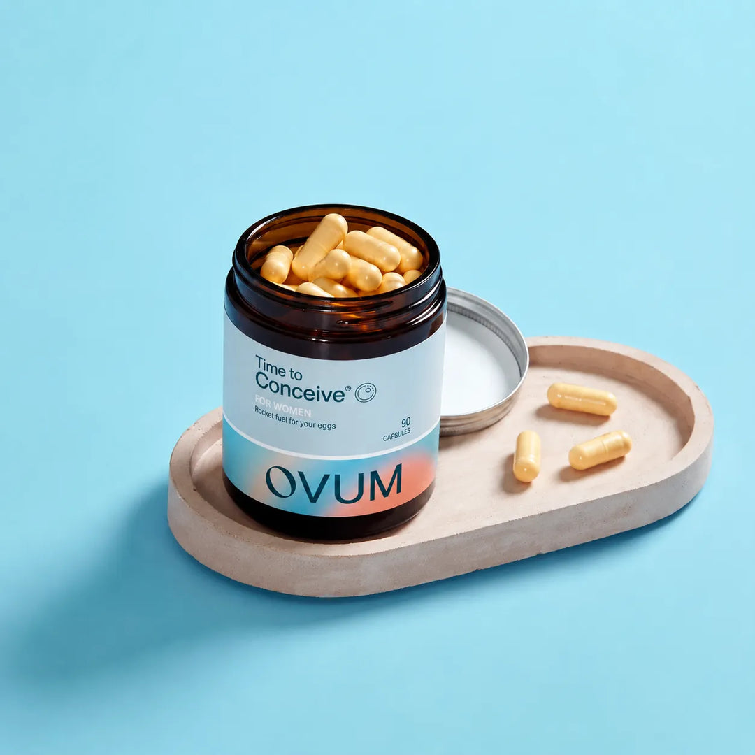 An open amber supplement bottle labeled 'Time to Conceive' and 'OVUM' sits on a light wood tray with several yellow capsules beside it, set against a solid light blue background.