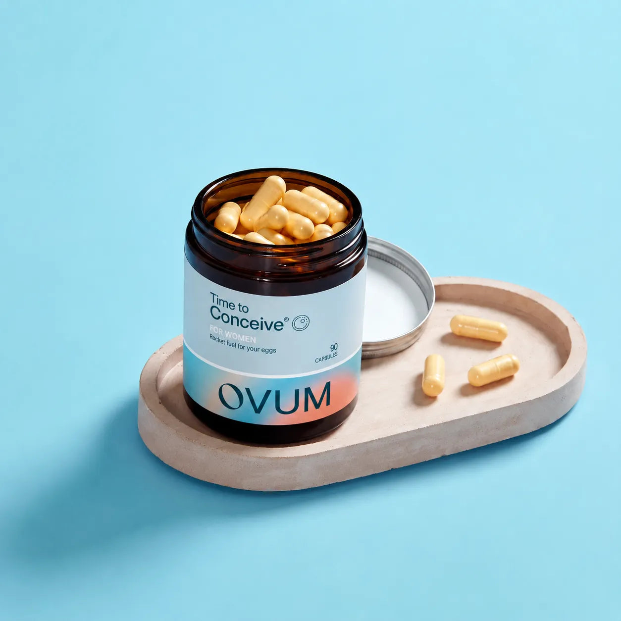An open amber supplement bottle labeled 'Time to Conceive' and 'OVUM' sits on a light wood tray with several yellow capsules beside it, set against a solid light blue background.