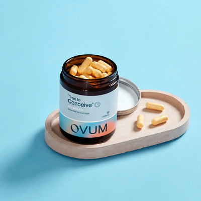 An open amber supplement bottle labeled 'Time to Conceive' and 'OVUM' sits on a light wood tray with several yellow capsules beside it, set against a solid light blue background.