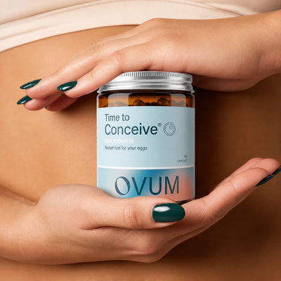 A close-up of a pregnant woman's hands holding an amber glass bottle of OVUM 'Time to Conceive' capsules against her stomach.