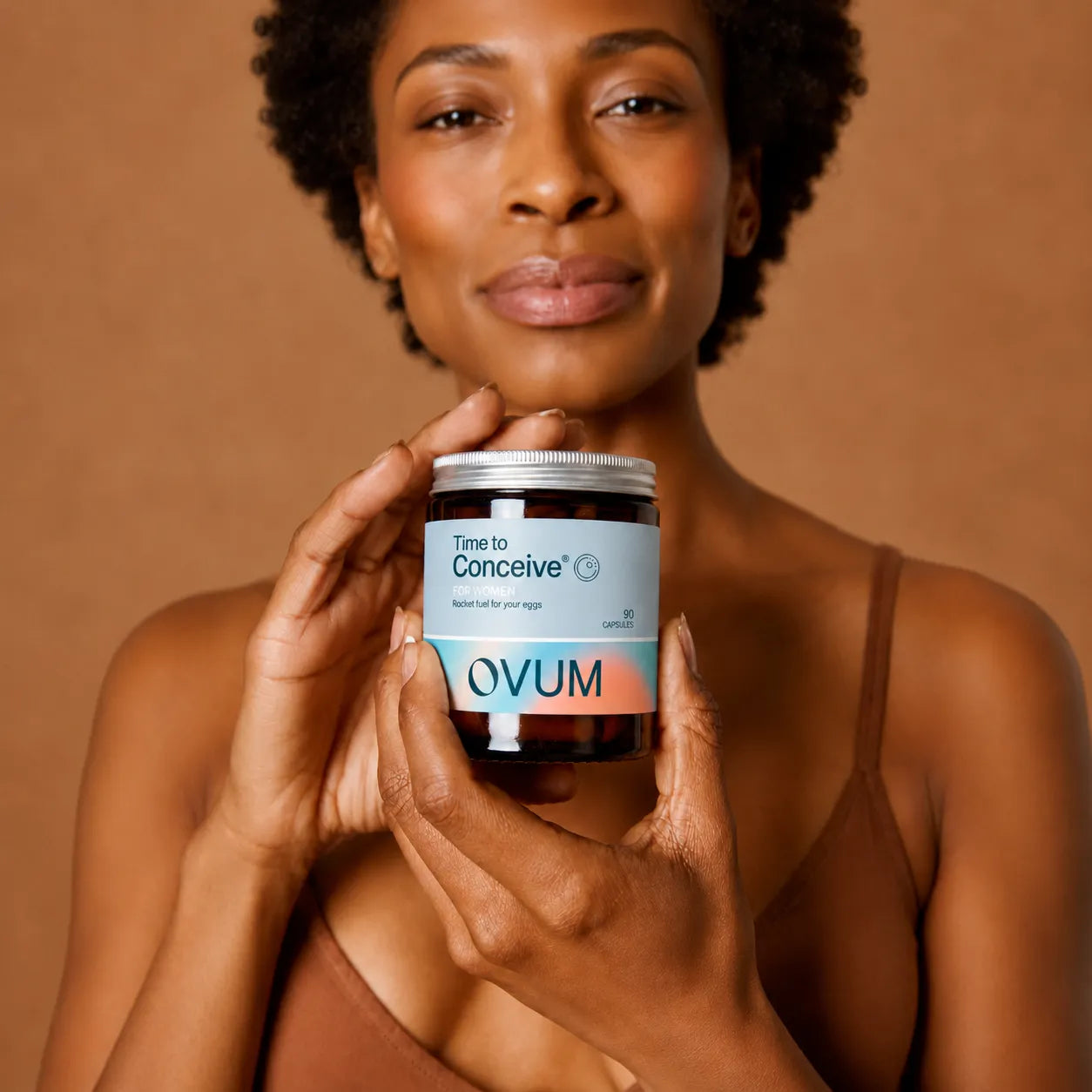 A woman holding a jar of OVUM Time to Conceive supplements against a warm brown studio background.