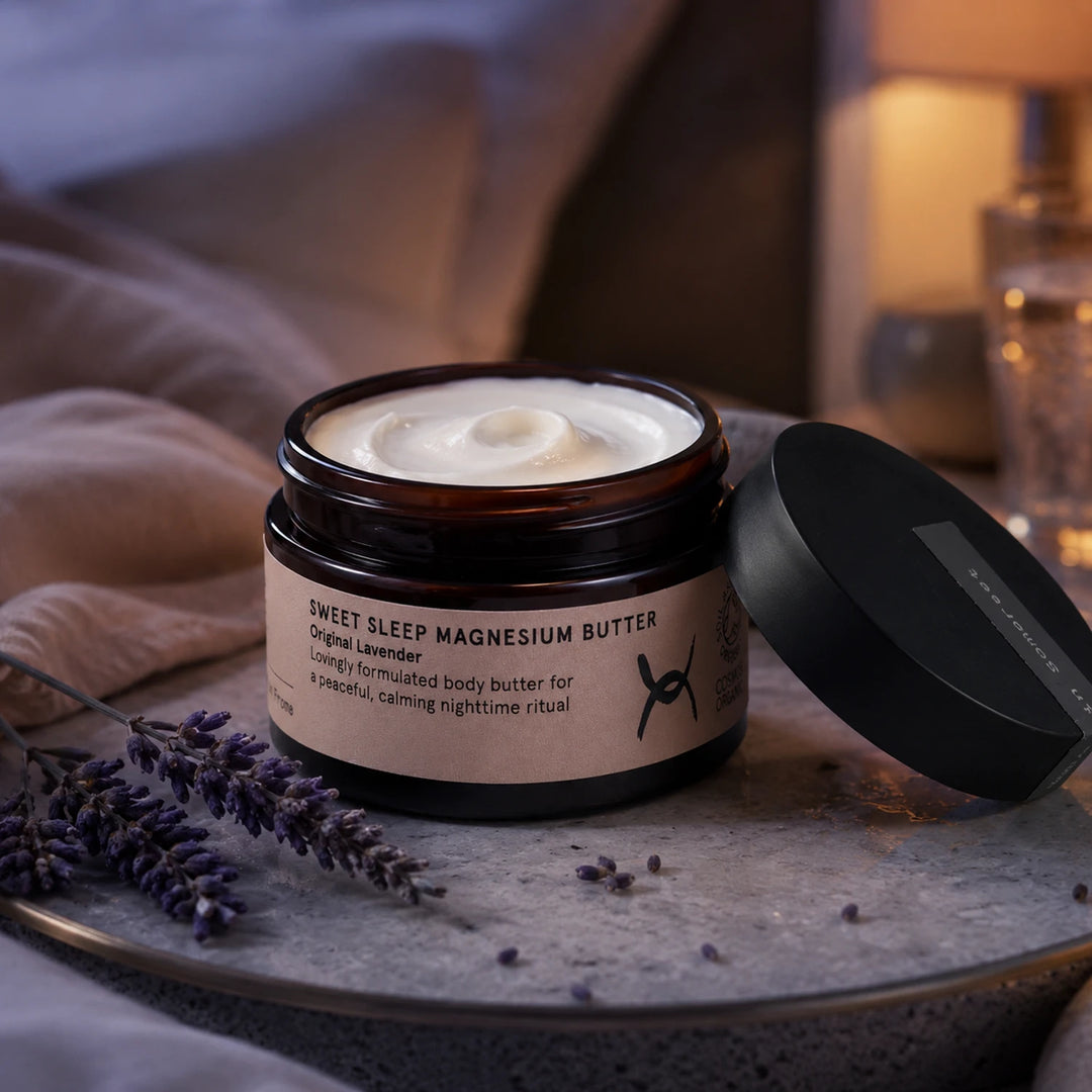 A jar of Sweet Sleep Magnesium Butter with its lid off, showing the white cream inside. The jar is placed on a gray stone tray with lavender sprigs, set against a cozy, softly lit bedroom background.