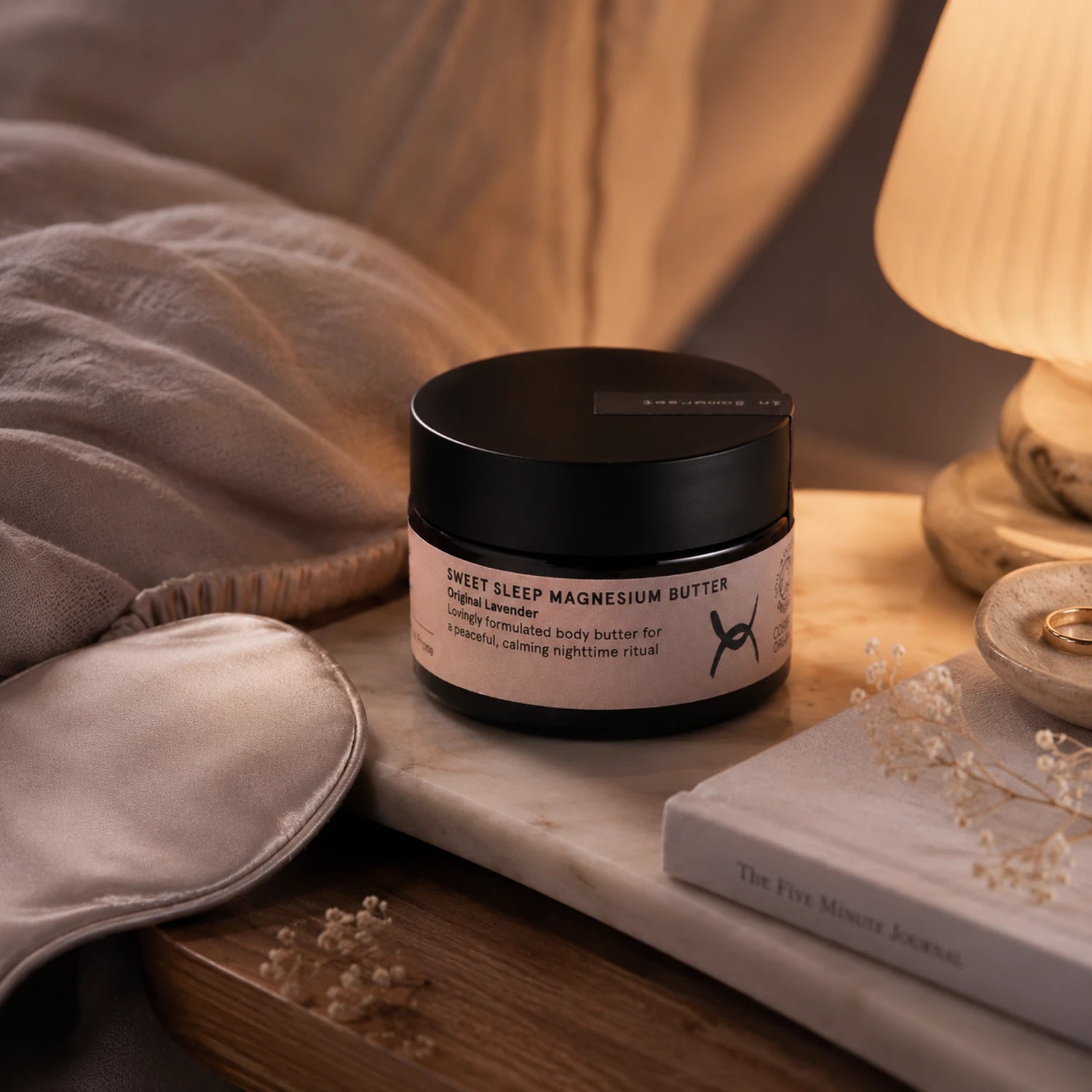A jar of Sweet Sleep Magnesium Butter sits on a bedside table next to a satin sleep mask, a lit lamp, and a book in a warm, cozy bedroom setting.