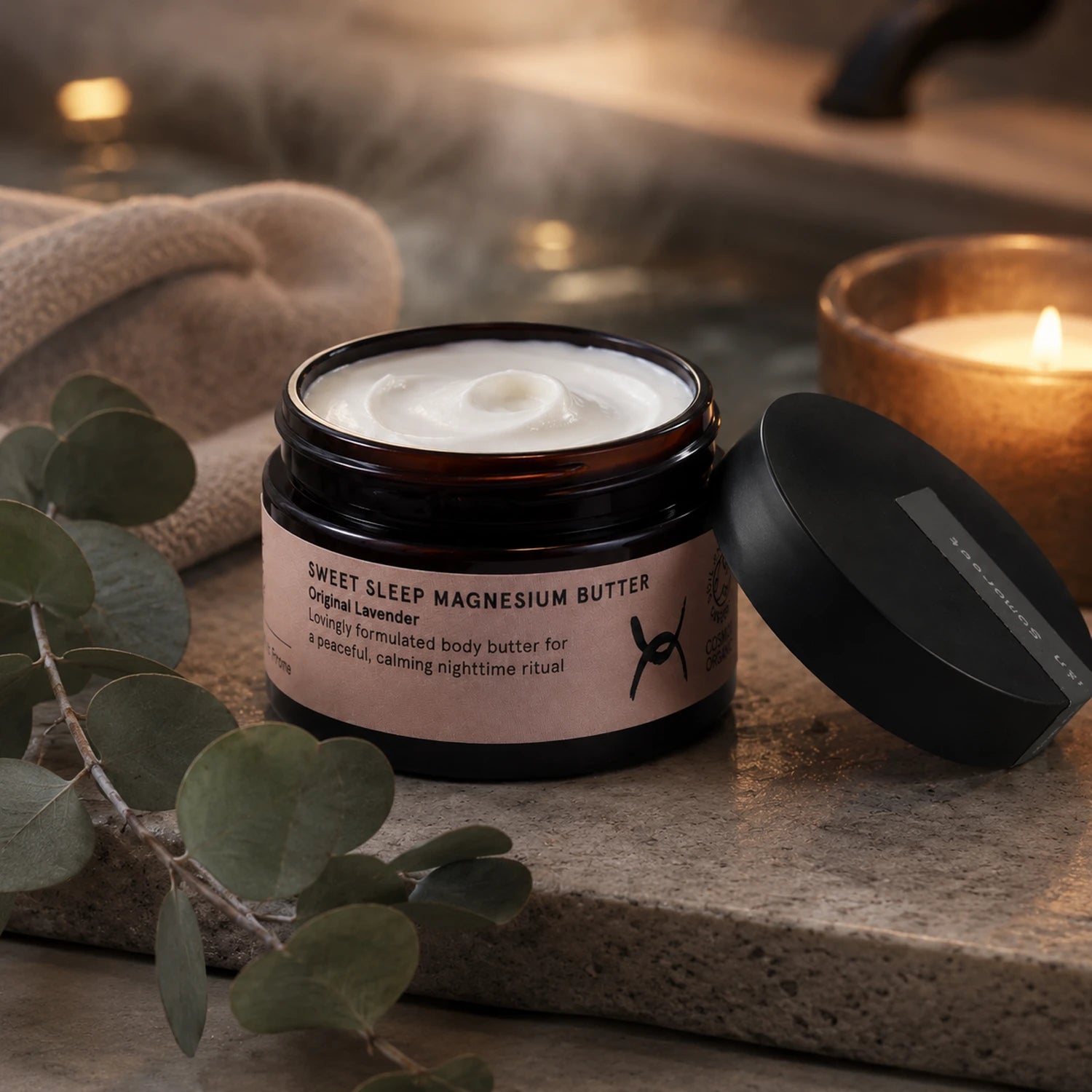 A jar of Sweet Sleep Magnesium Butter with its lid off, showing the white cream inside. The jar is placed on a stone surface next to a towel, eucalyptus leaves, and a lit candle in a warm, relaxing setting.