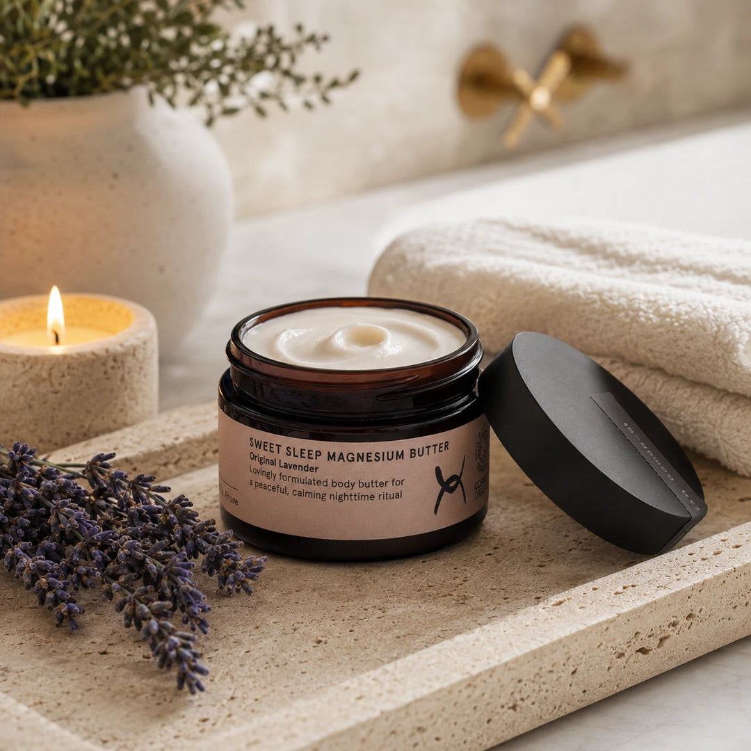 An open jar of Sweet Sleep Magnesium Butter with its black lid resting beside it on a stone tray, styled with lavender sprigs, a lit candle, folded towels, and a blurred bathroom background.