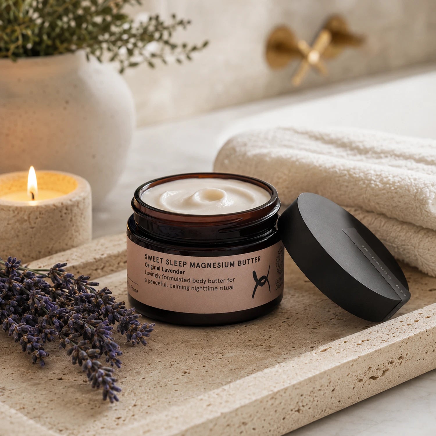 An open jar of Sweet Sleep Magnesium Butter with its black lid resting beside it on a stone tray, styled with lavender sprigs, a lit candle, folded towels, and a blurred bathroom background.