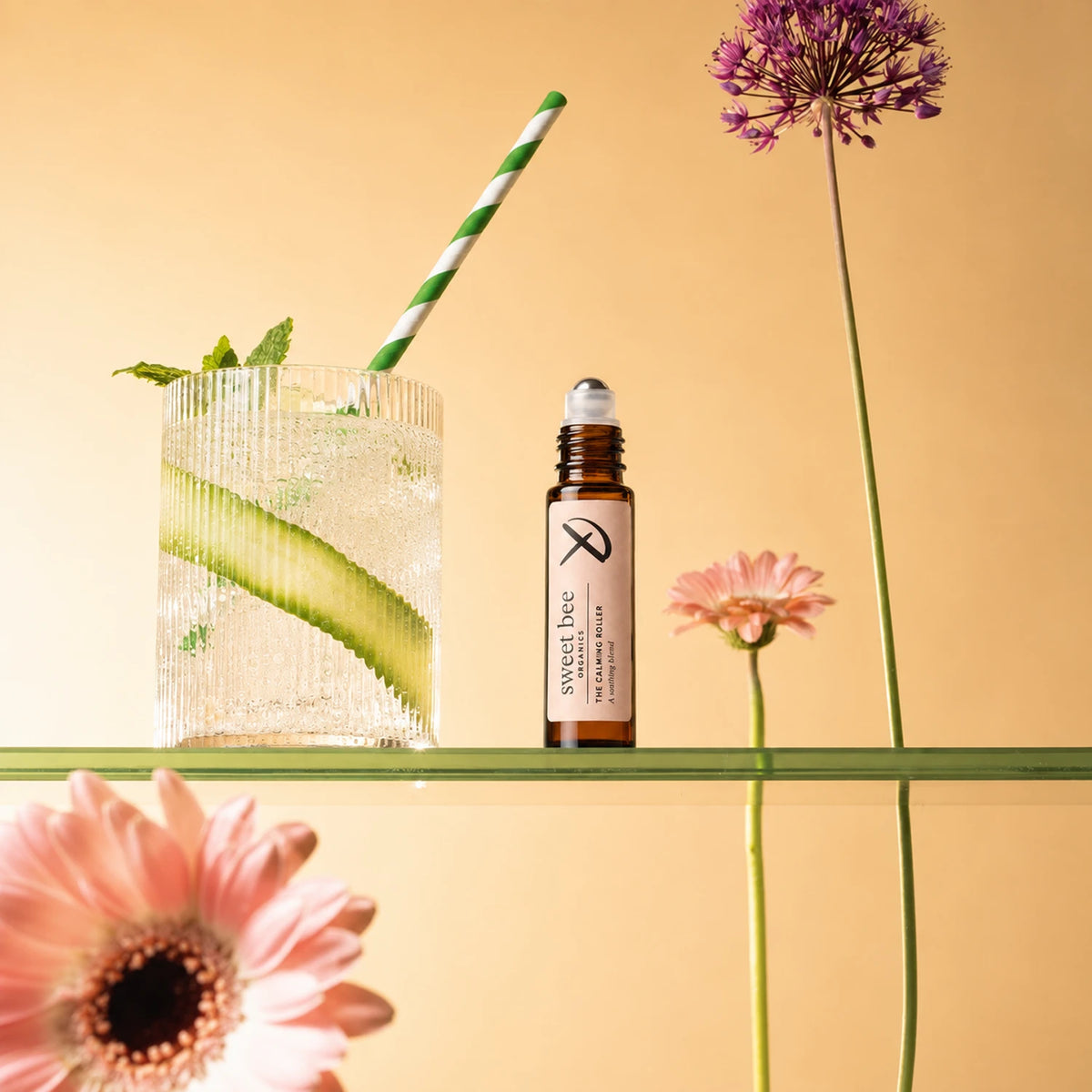 A small amber bottle labeled Sweet Bee essential oil sits on a clear glass shelf beside a ribbed glass drink with a green-and-white striped straw, cucumber slice, mint, and flowers against a warm beige background.