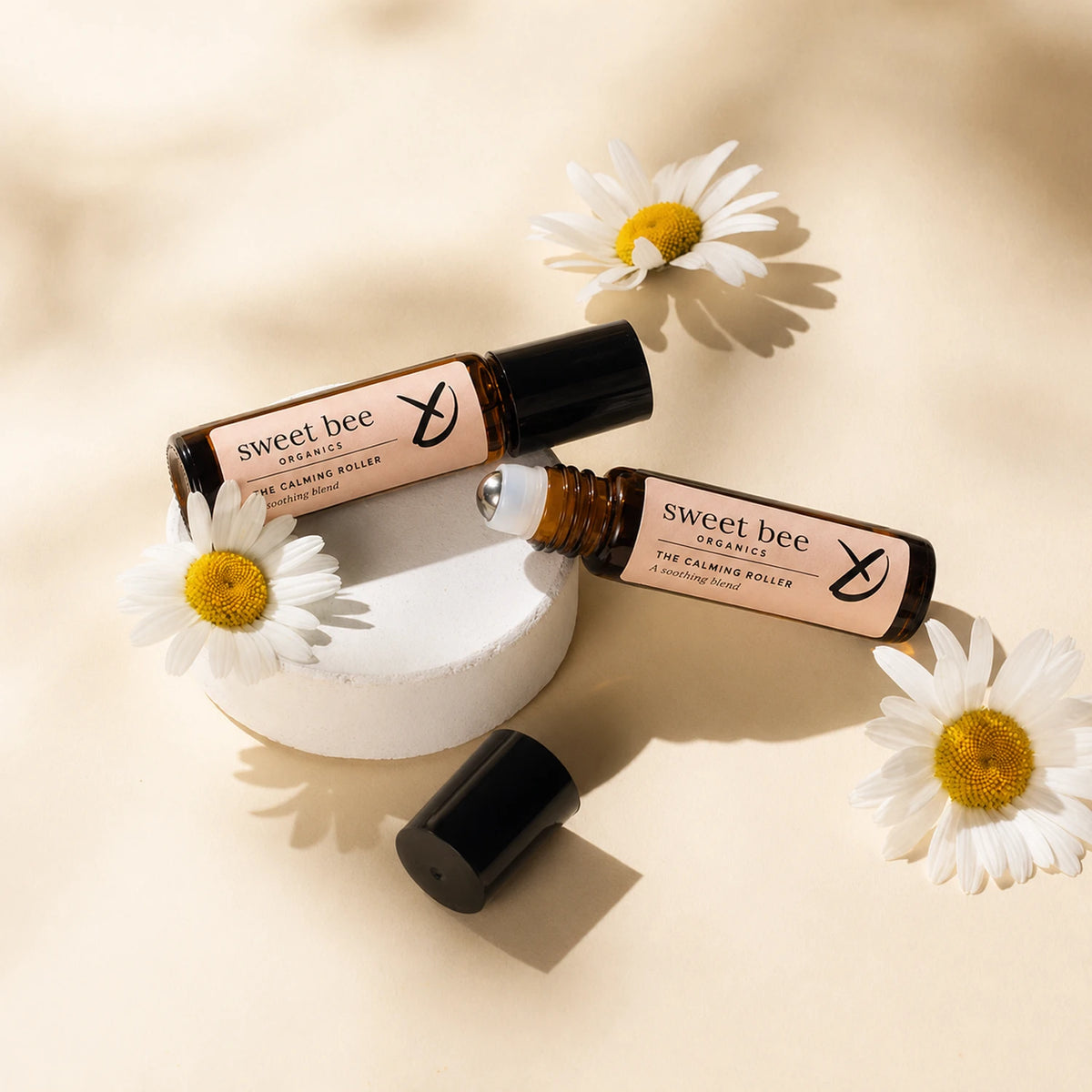 Two amber glass roll-on bottles labeled 'sweet bee' are arranged on a warm beige surface with white daisy flowers and a small white pedestal. One bottle is open with its black cap placed beside it.