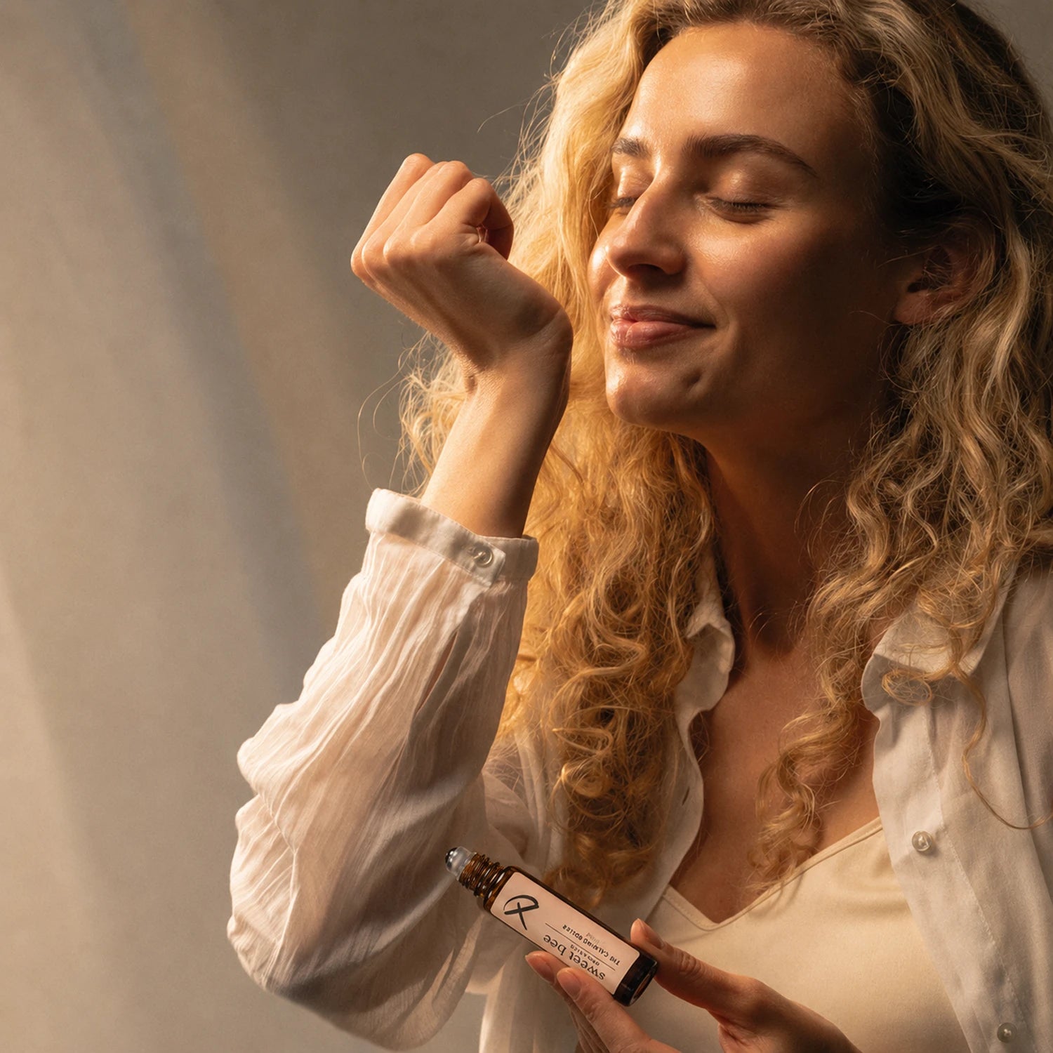 A woman with long curly blonde hair holds a small amber roller bottle near her face, appearing to smell or inhale from it. She is wearing a white button-up shirt over a cream top against a softly lit neutral background.
