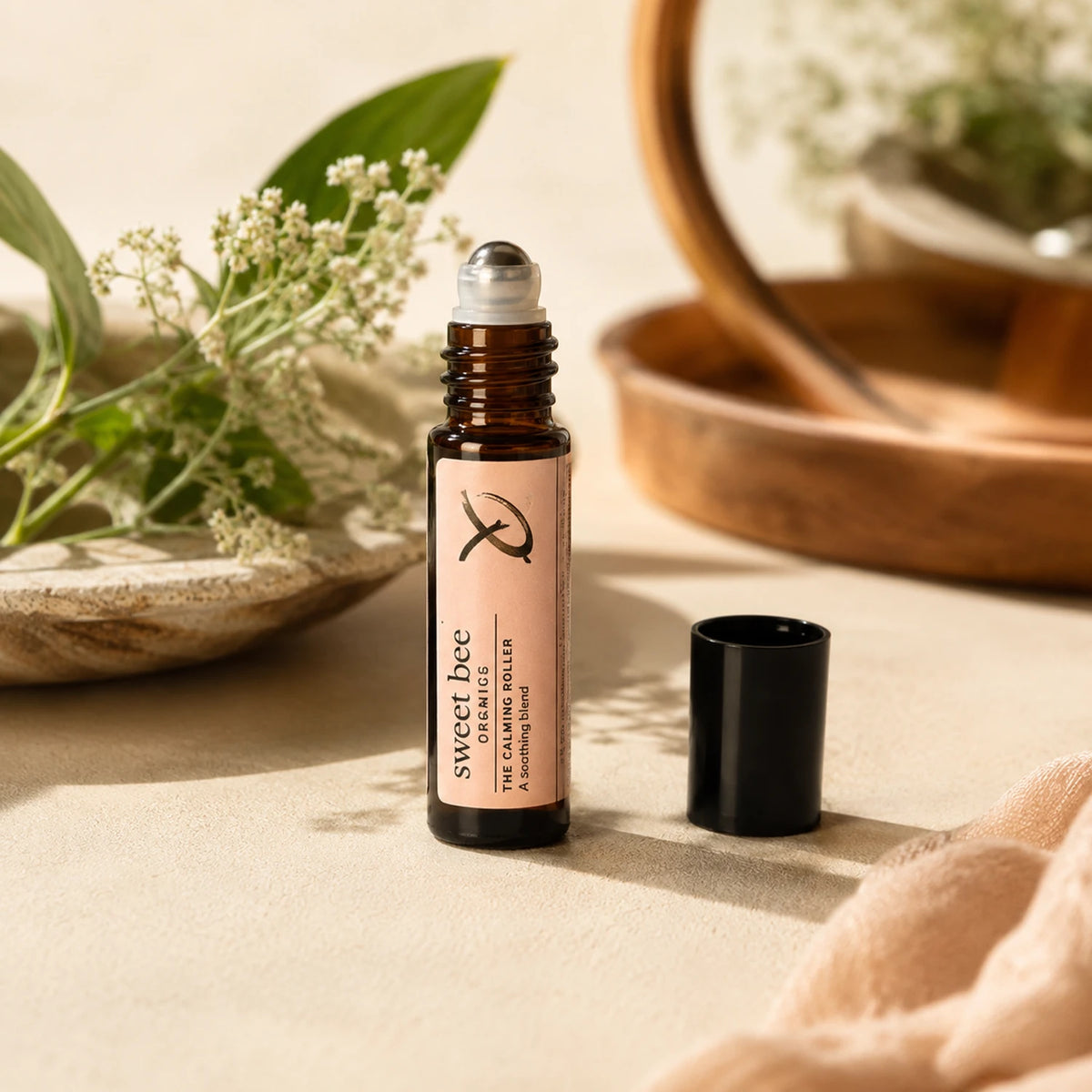 A small amber glass roller bottle labeled 'sweet bee botanicals' stands on a beige surface with its black cap beside it. The scene includes soft-focus greenery, a wooden bowl, and a mirror in the background, creating a warm natural setting.